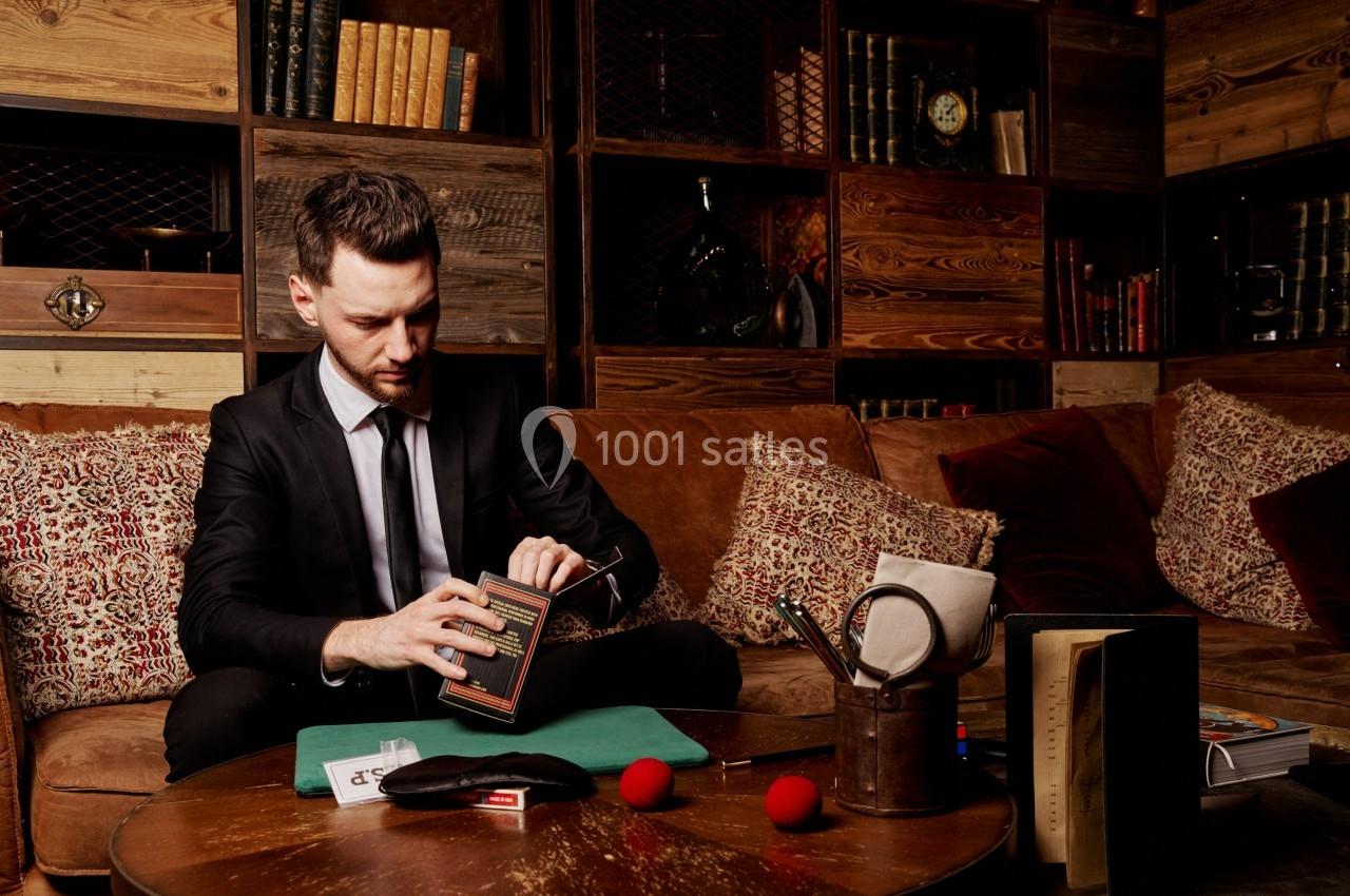 Un homme en costume assis dans un salon cosy, manipulant un livre entouré de documents et d'objets divers.