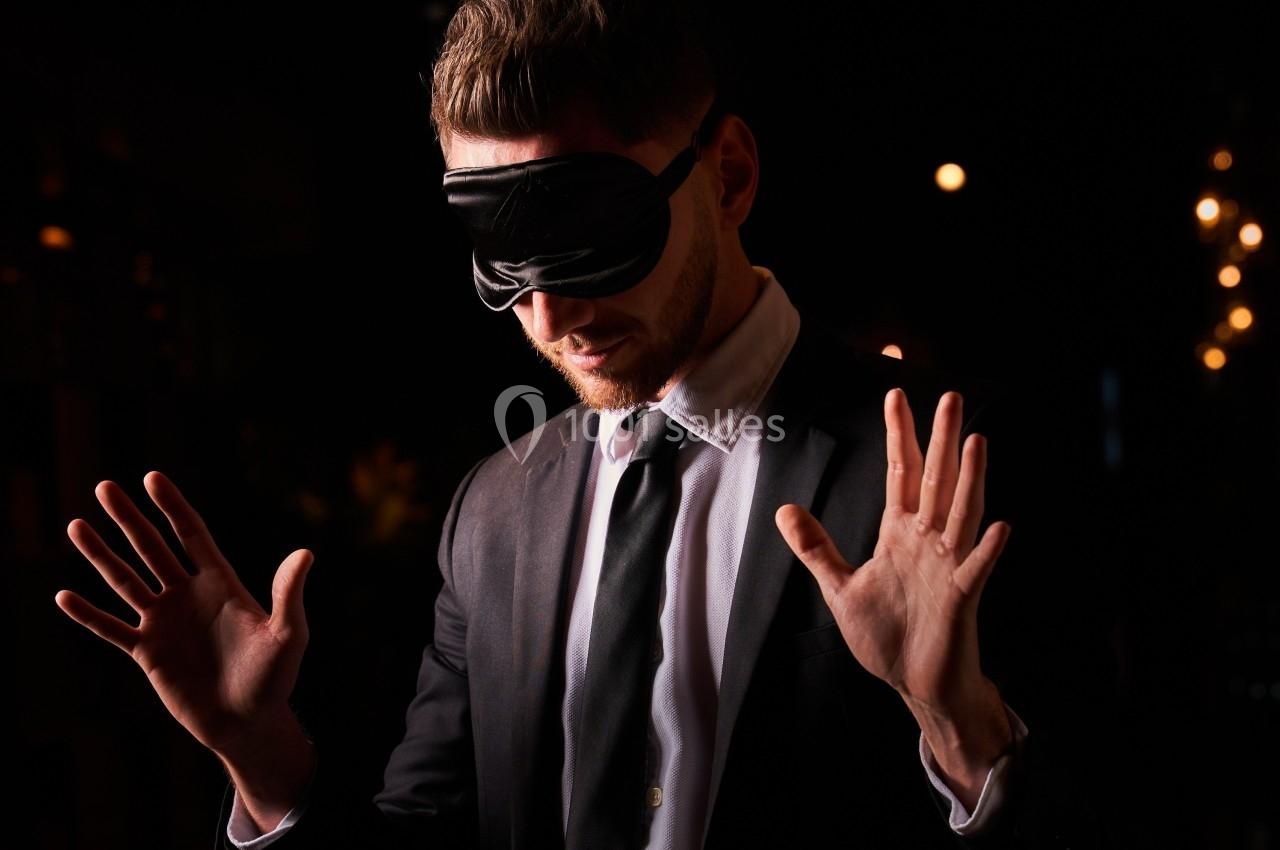 Un homme en costume, les mains levées, portant un masque noir sur les yeux dans un environnement sombre.