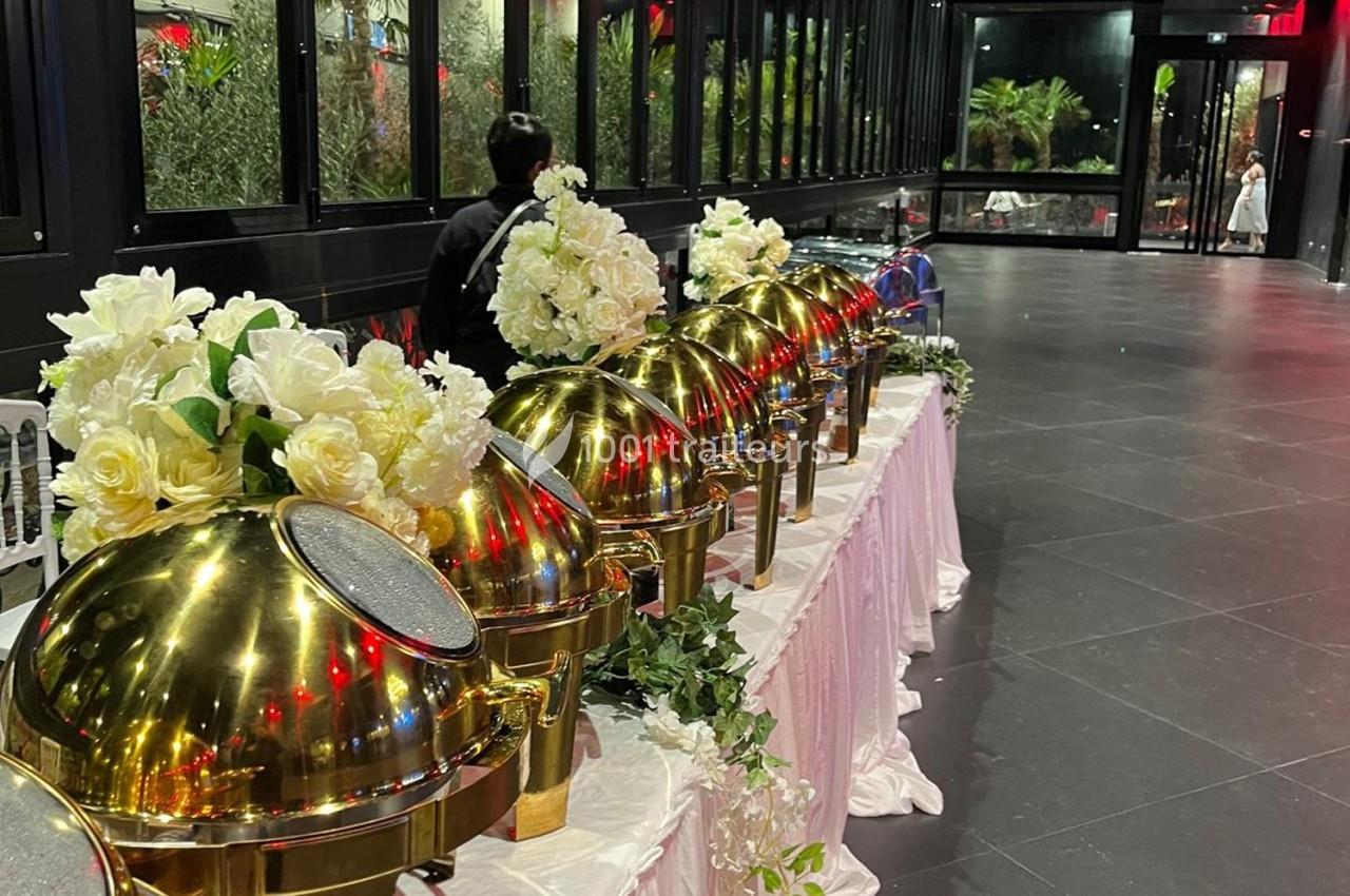 Buffet avec plats sous cloches dorées, décoré de fleurs blanches, dans une salle vitrée avec vue sur l'extérieur.