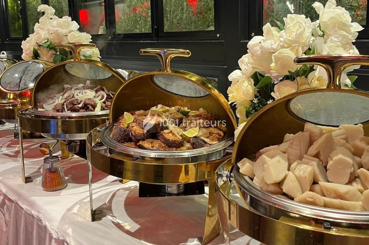 Buffet avec plats variés sous cloches dorées, incluant viandes grillées, légumes et décorations florales blanches.