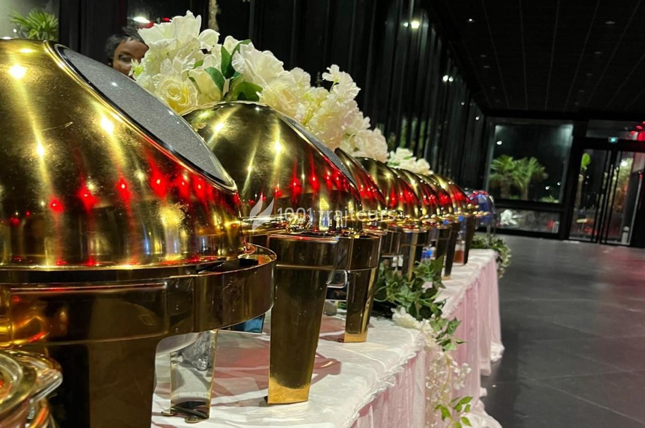 Rangée de plats en métal doré sur une table décorée de fleurs blanches dans une salle sombre.
