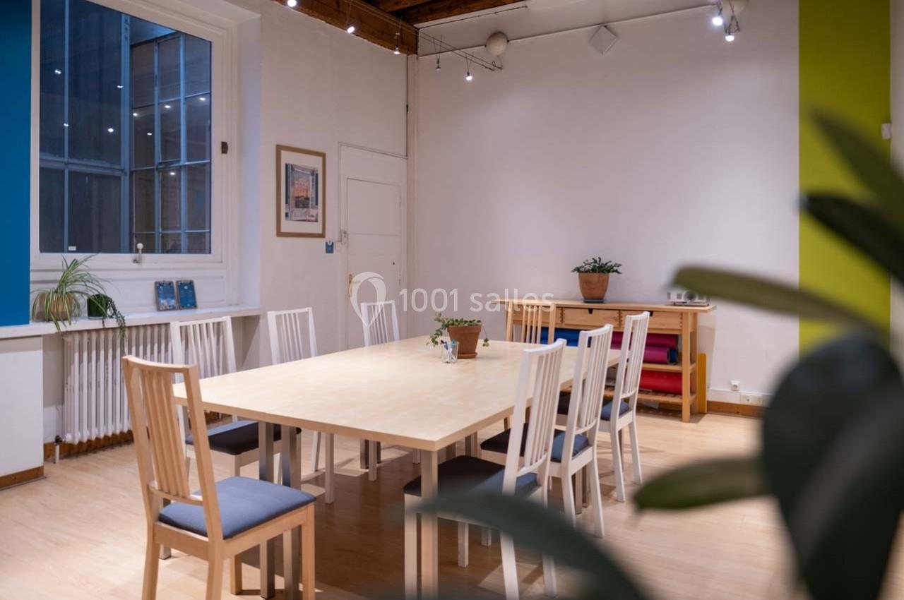 Salle lumineuse avec une grande table en bois entourée de chaises, des plantes et des étagères en arrière-plan.