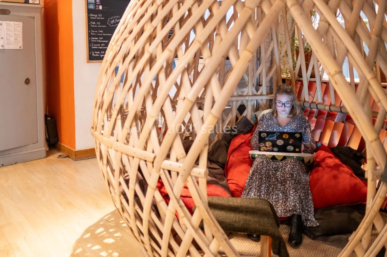 Une femme travaille sur un ordinateur portable, assise dans une structure en bois tressé avec des coussins colorés.
