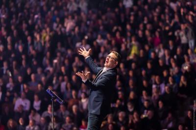 Une main verse du vin blanc dans un verre transparent, avec des bouteilles en arrière-plan flou.