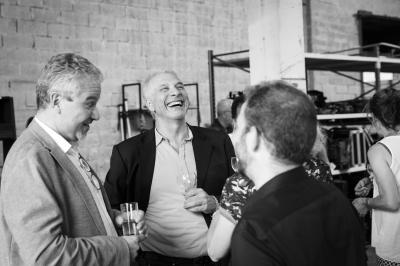 Une main verse du vin blanc dans un verre transparent, avec des bouteilles en arrière-plan flou.