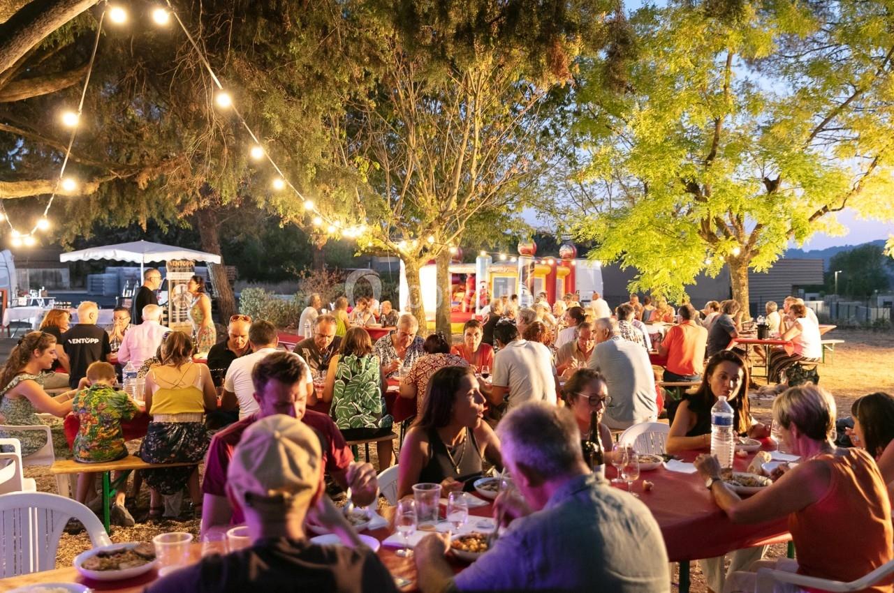 Des personnes dînent en plein air sous des guirlandes lumineuses, entourées d'arbres et de stands de nourriture.