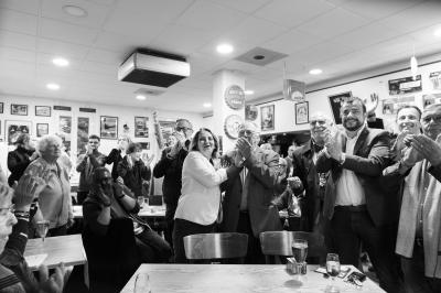 Une main verse du vin blanc dans un verre transparent, avec des bouteilles en arrière-plan flou.