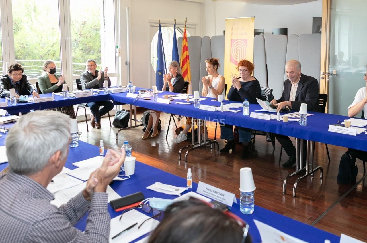 Des personnes assises autour d'une table en réunion, avec des documents, drapeaux et panneaux en arrière-plan.