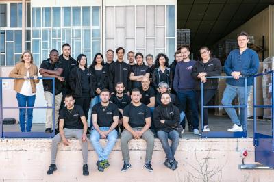 Une main verse du vin blanc dans un verre transparent, avec des bouteilles en arrière-plan flou.