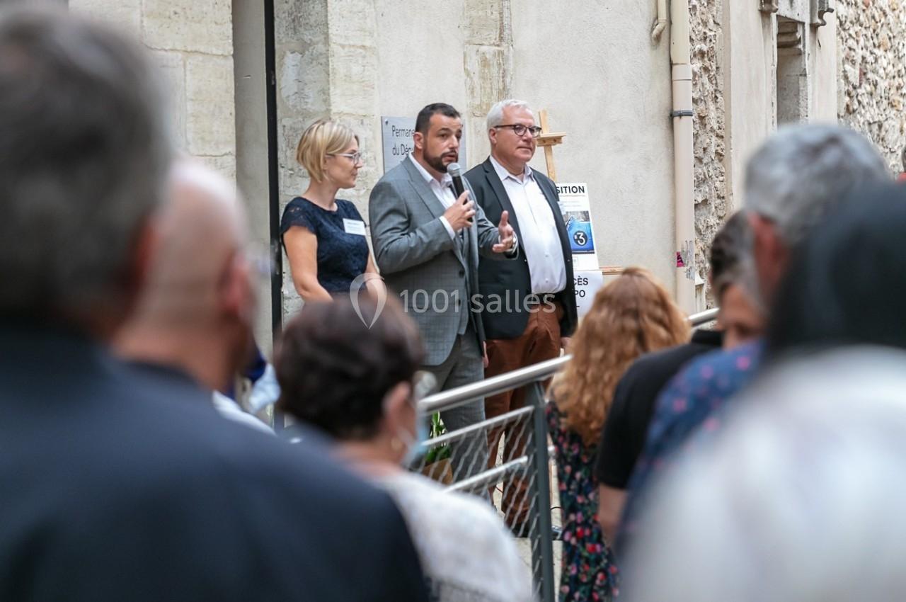 Un homme s'exprime au micro devant un public rassemblé, accompagné de deux personnes, près d'un bâtiment ancien.