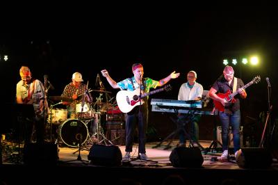 Une main verse du vin blanc dans un verre transparent, avec des bouteilles en arrière-plan flou.