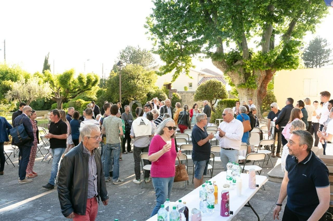 Groupe de personnes rassemblées en extérieur autour de tables avec boissons, dans un cadre arboré et ensoleillé.