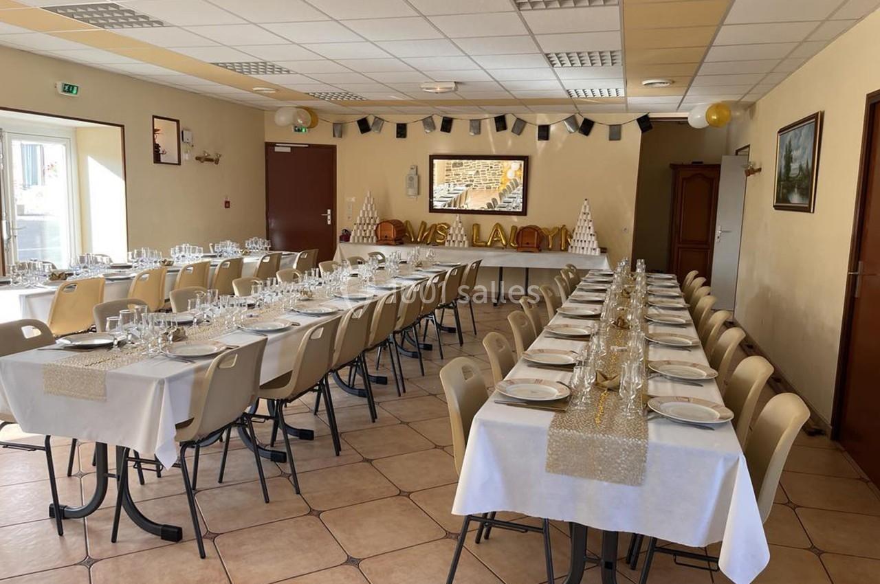 Salle décorée pour un événement, avec tables alignées, nappes blanches, vaisselle dressée et décorations festives.