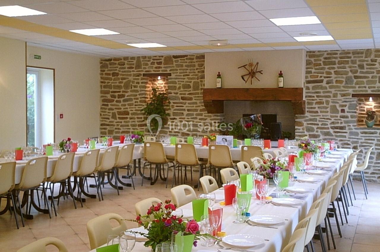 Salle de réception avec de longues tables dressées, décorée de fleurs et de couleurs vives, murs en pierre apparente.