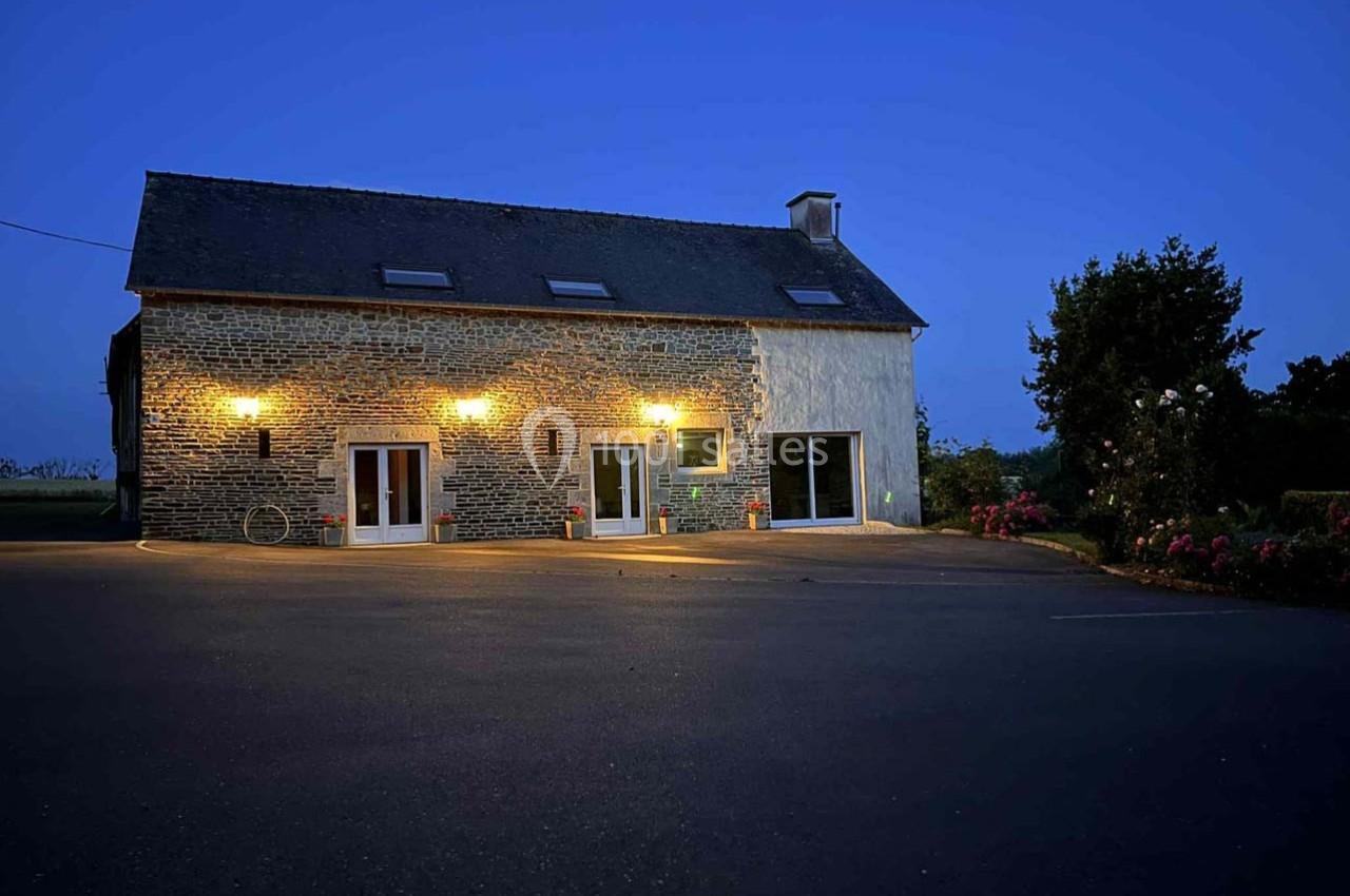 Façade d'une maison en pierre éclairée par des lumières extérieures, avec une cour asphaltée au crépuscule.