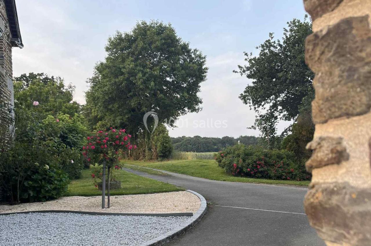 Allée bordée de pelouse et de rosiers, menant à un paysage champêtre avec des arbres sous un ciel dégagé.