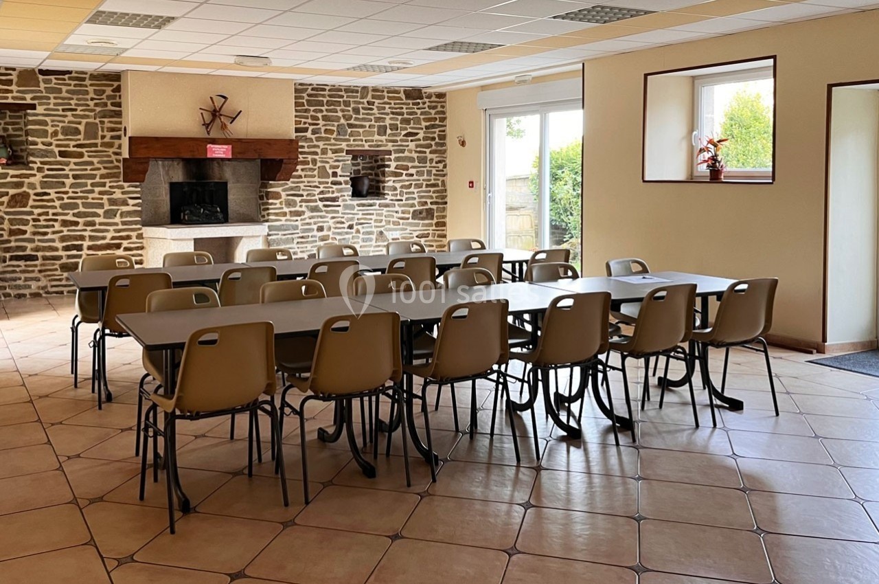 Salle lumineuse avec murs en pierre, cheminée, tables et chaises alignées, donnant sur des fenêtres et une porte vitrée.