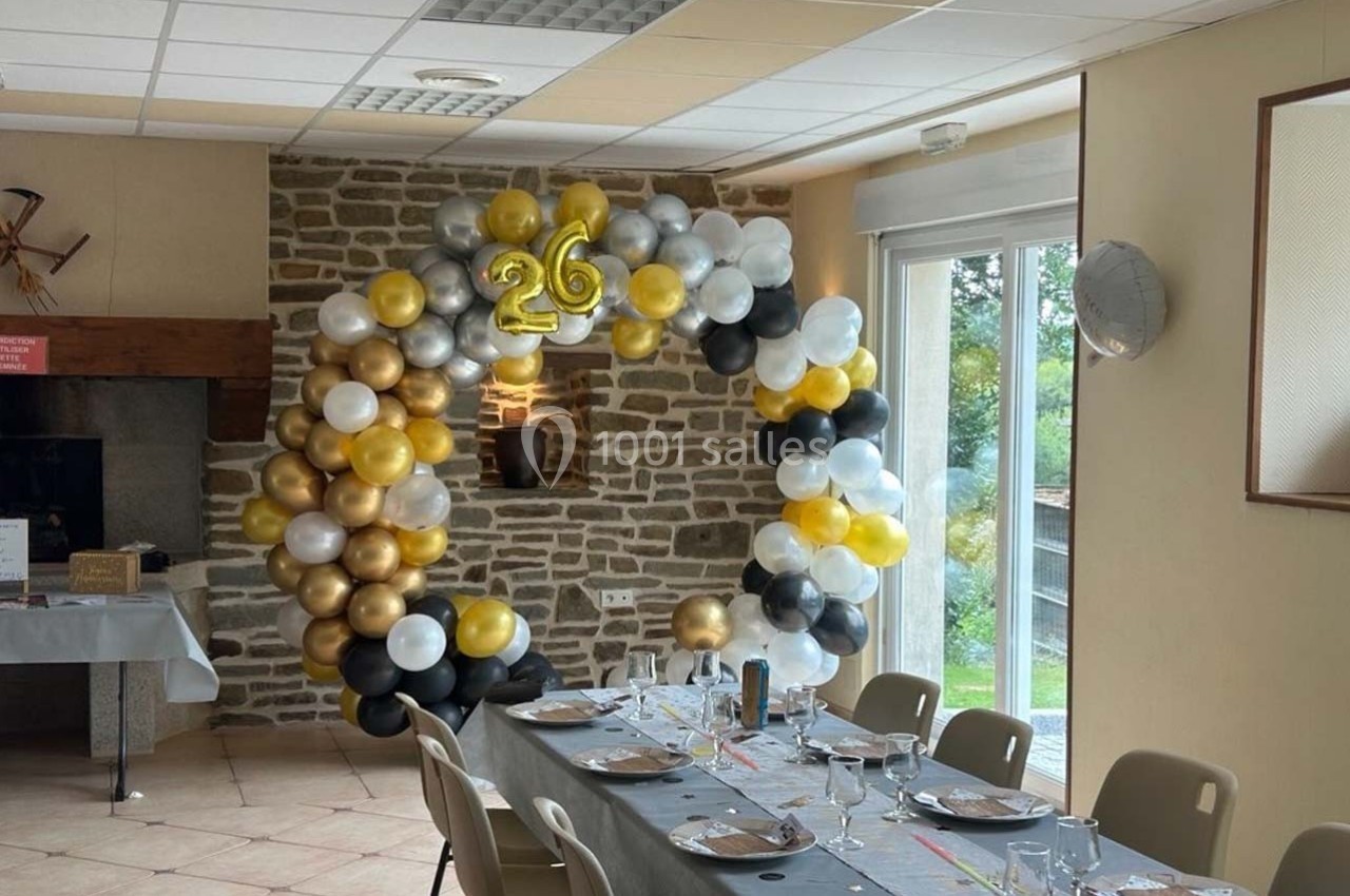 Arc de ballons dorés, blancs, noirs et argentés formant le chiffre 26, décorant une salle avec table dressée.