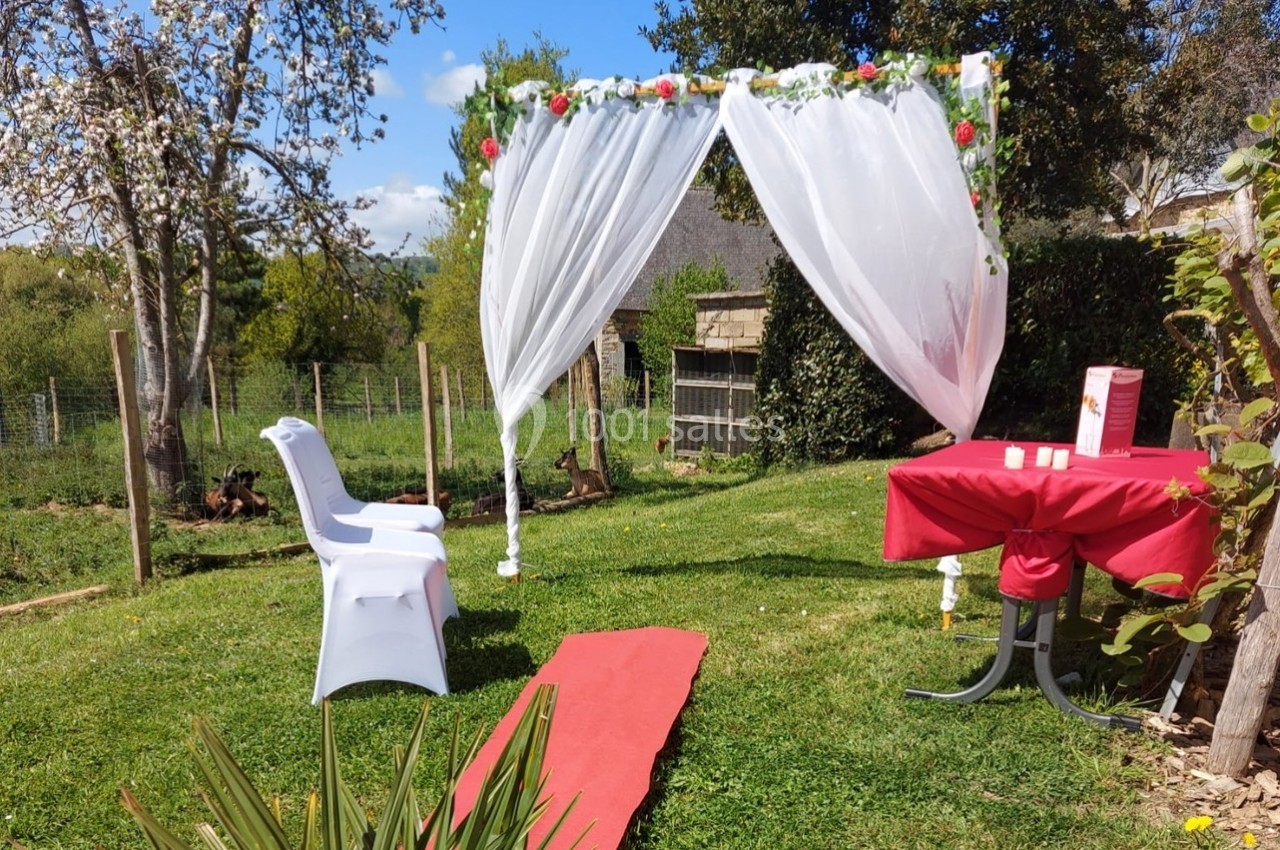 Arche décorée de voilages et de fleurs dans un jardin, avec chaise blanche, table rouge et allée en tapis rouge.