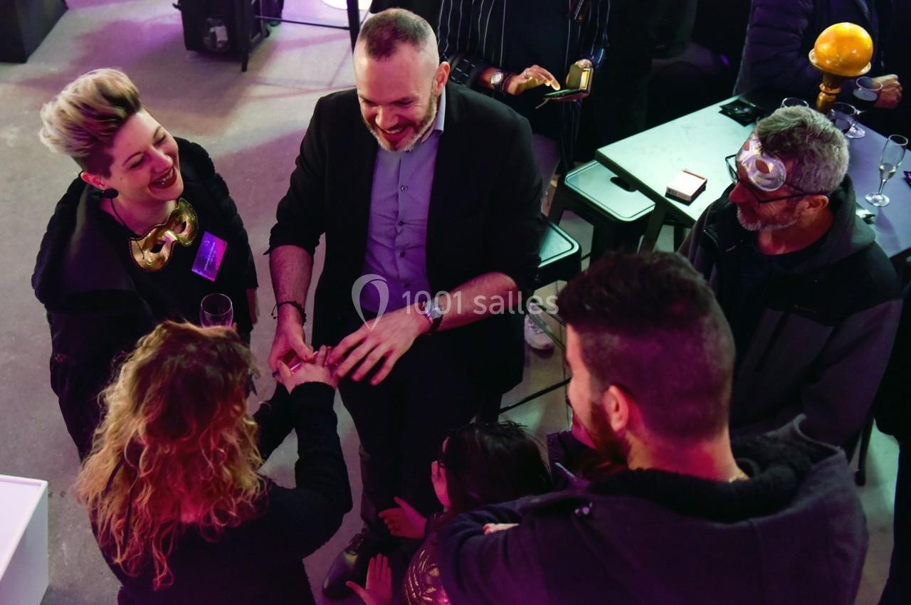 Un magicien réalise un tour de cartes entouré de spectateurs souriants dans un espace intérieur animé.