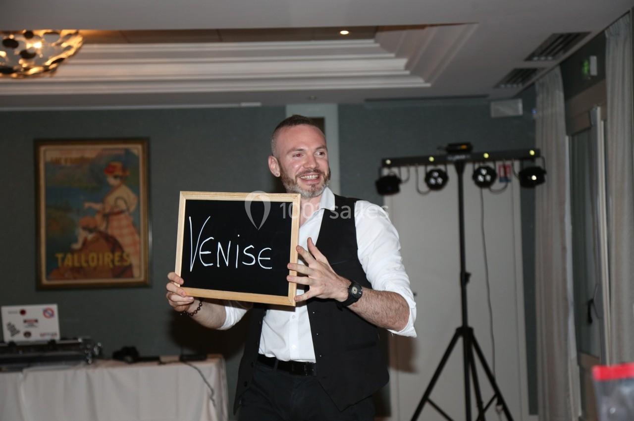 Un homme souriant tient une ardoise avec le mot ’Venise’ dans une salle décorée.
