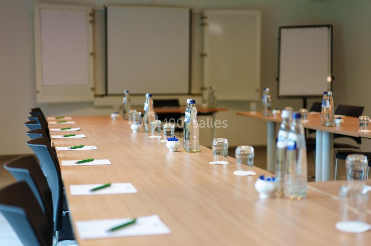 Salle de réunion avec tables disposées en U, contenant des bouteilles d'eau, des verres, des stylos et des blocs-notes.