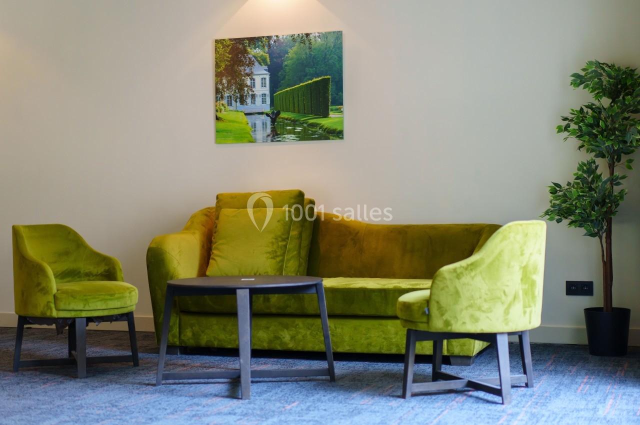 Salon avec canapé et fauteuils verts, table basse ronde en bois, plante en pot et tableau mural représentant un jardin.
