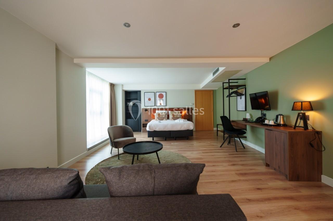 Chambre spacieuse avec lit double, coin salon, bureau, télévision et décoration moderne dans des tons neutres.