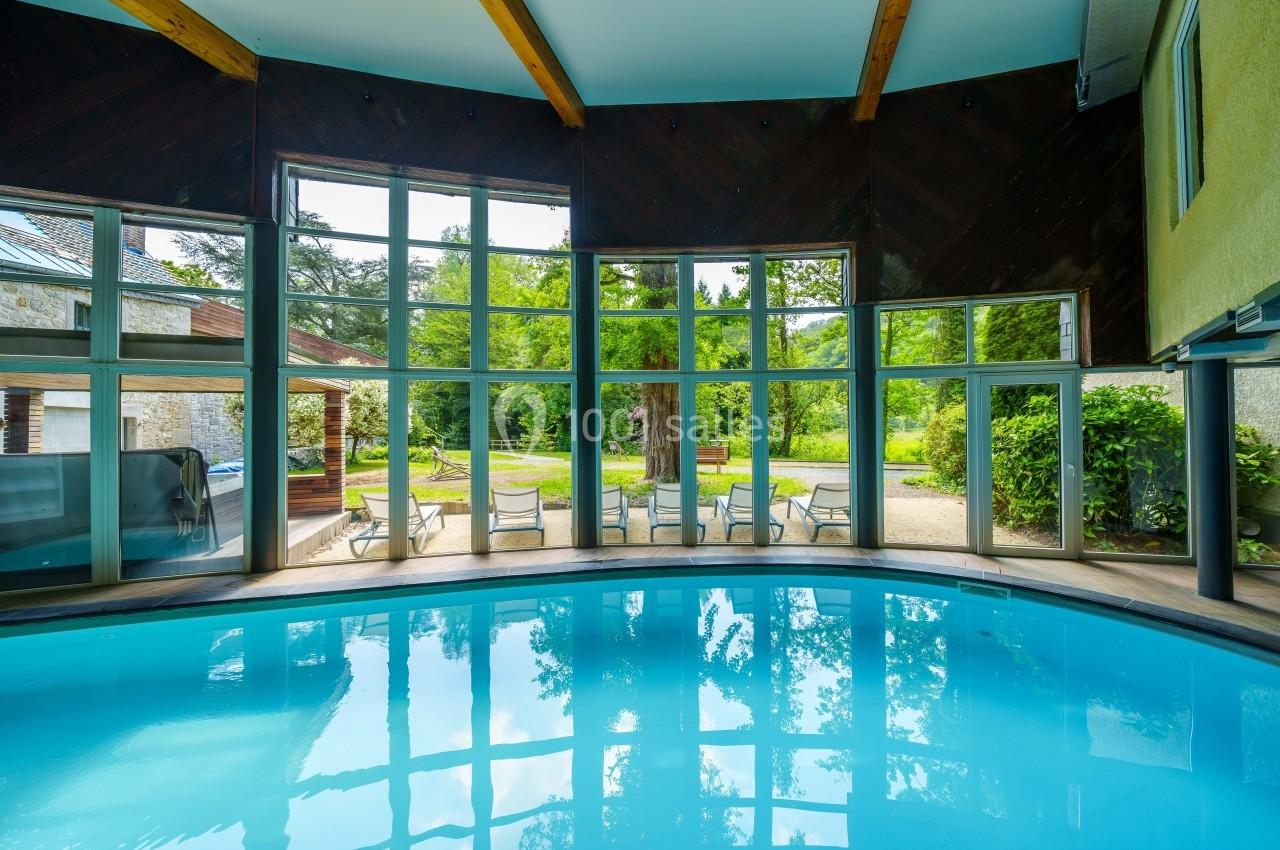 Piscine intérieure avec grandes baies vitrées offrant une vue sur un jardin verdoyant et des chaises disposées à l'extérieur.