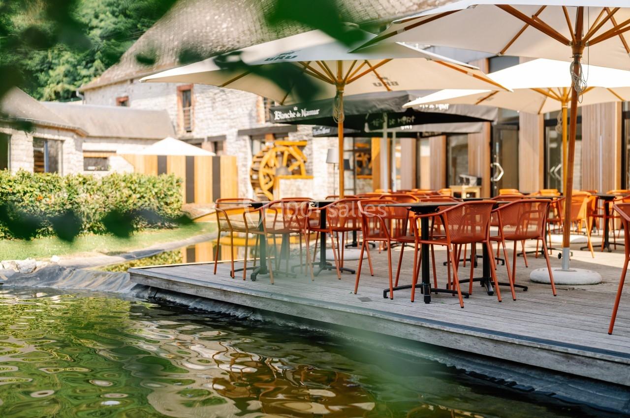 Terrasse avec tables et chaises orange sous des parasols, située près d'un étang et entourée de verdure.