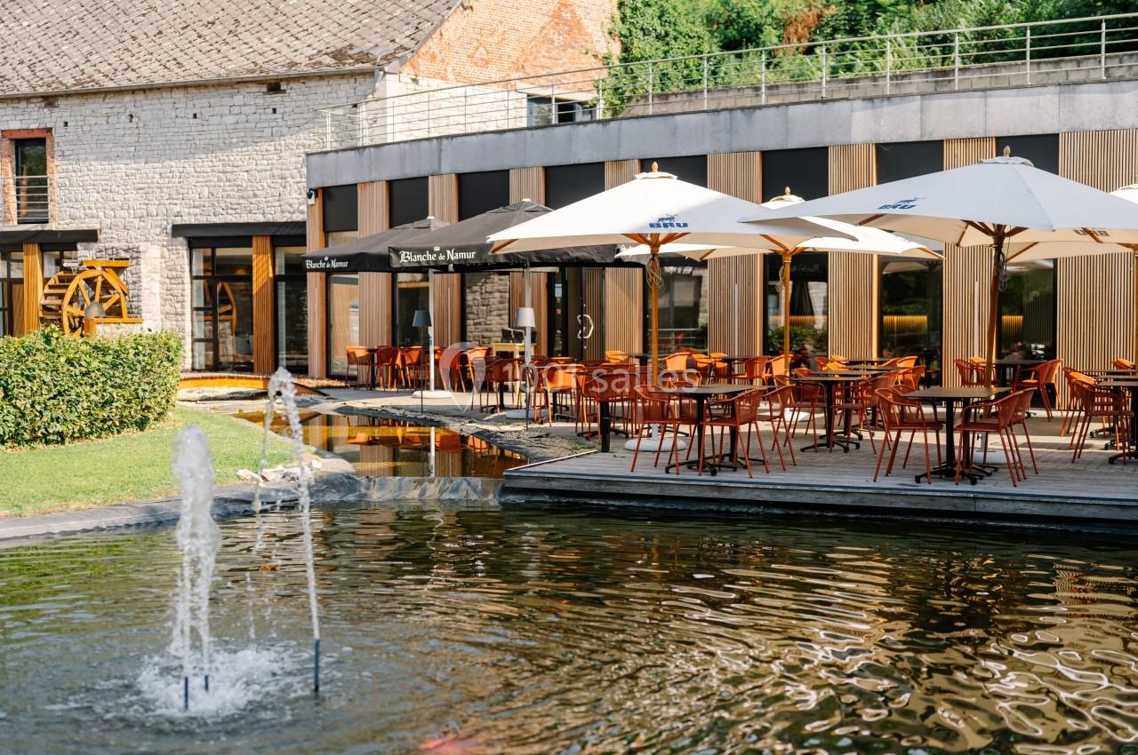 Terrasse d'un restaurant avec tables, chaises et parasols, bordée d'un bassin avec une fontaine, dans un cadre verdoyant.