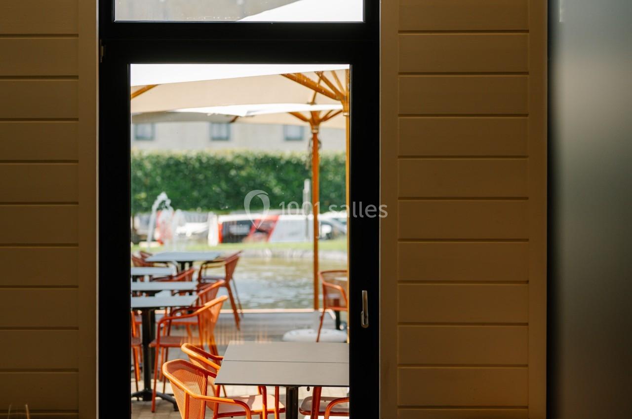 Vue depuis l'intérieur d'un restaurant sur une terrasse extérieure avec des tables, chaises orange et parasols.