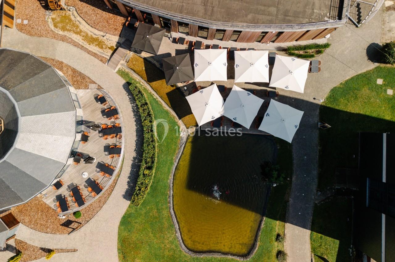 Vue aérienne d'une terrasse avec parasols blancs, entourée d'un bâtiment circulaire et d'un bassin avec végétation.
