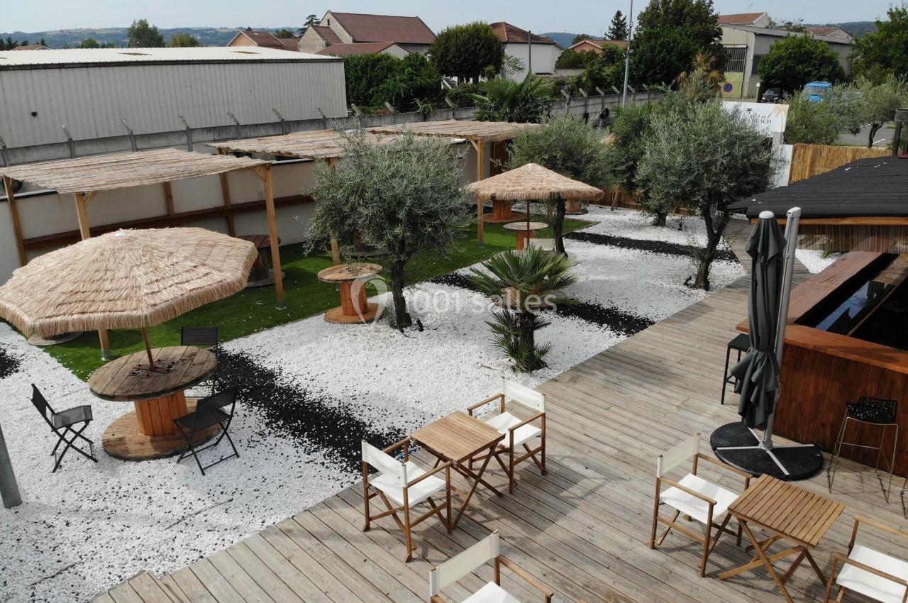 Terrasse extérieure avec tables, chaises, parasols en paille, oliviers et sol en gravier blanc, entourée de bâtiments.