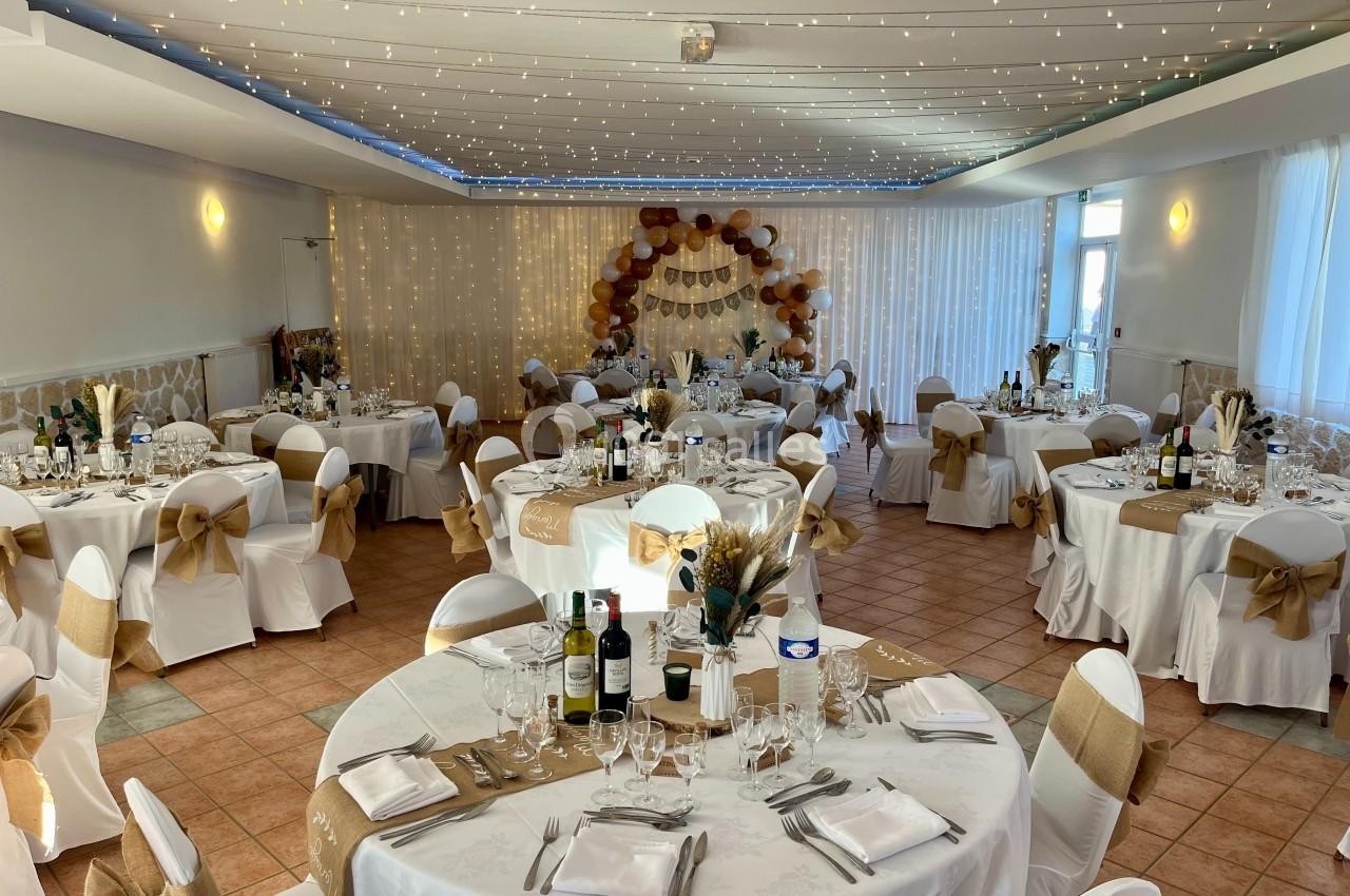 Salle de réception décorée avec des tables rondes, nappes blanches, chaises ornées de nœuds et guirlandes lumineuses au…