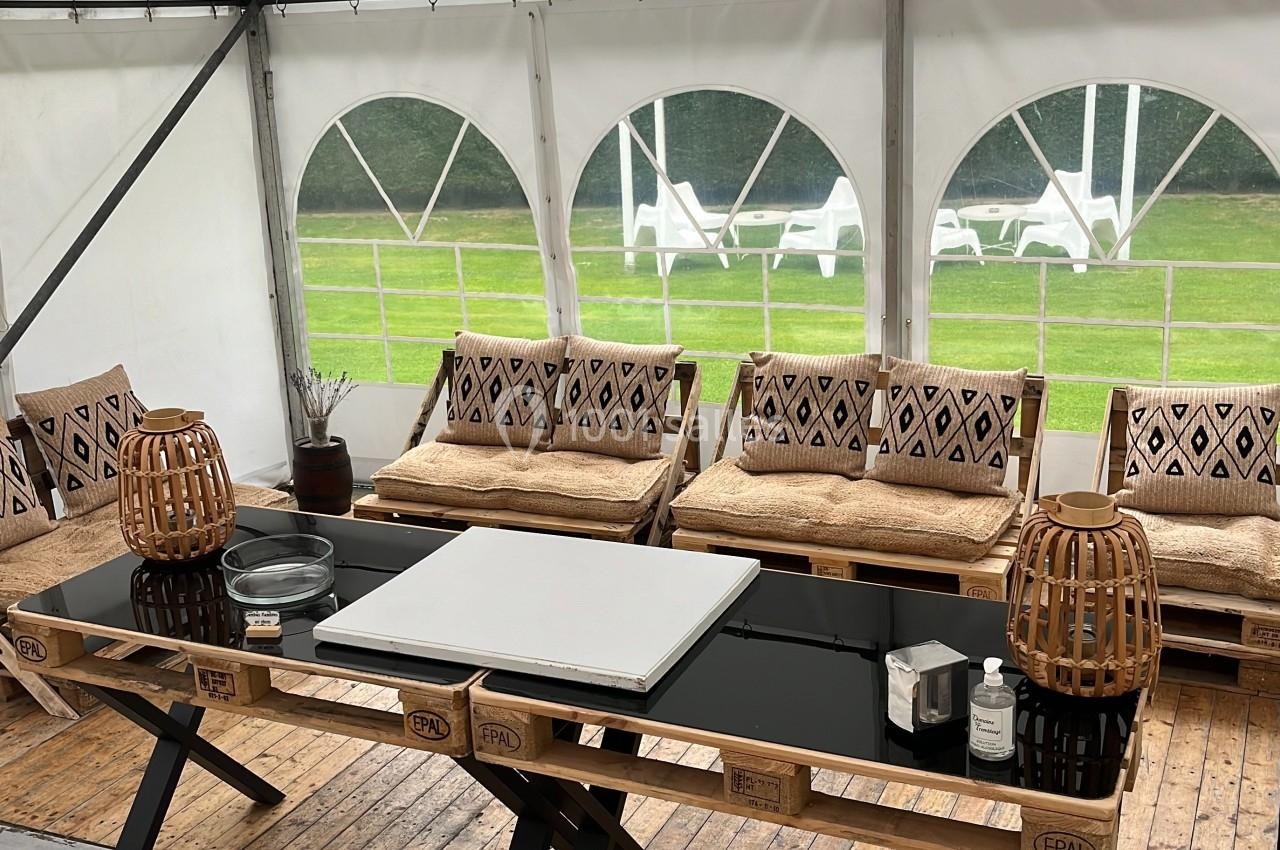 Table en bois avec plateau noir et objets décoratifs, entourée de bancs et fauteuils sous une tente ouverte sur un jardin.