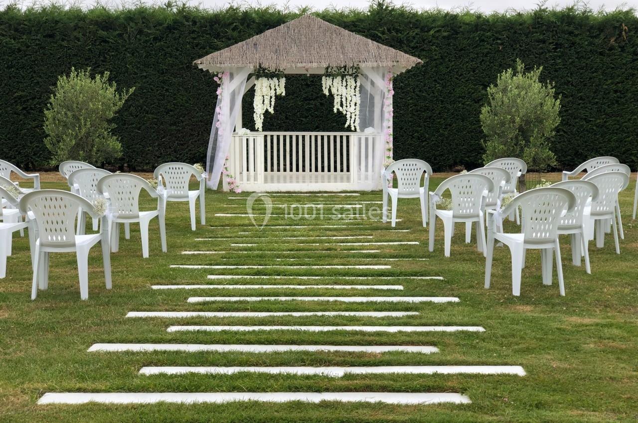 Allée en gazon bordée de chaises blanches menant à une pergola décorée pour une cérémonie en plein air.