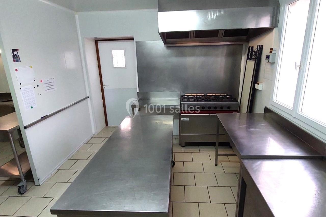 Cuisine professionnelle avec plans de travail en inox, cuisinière équipée d'une hotte et éclairage naturel par une fenêtre.