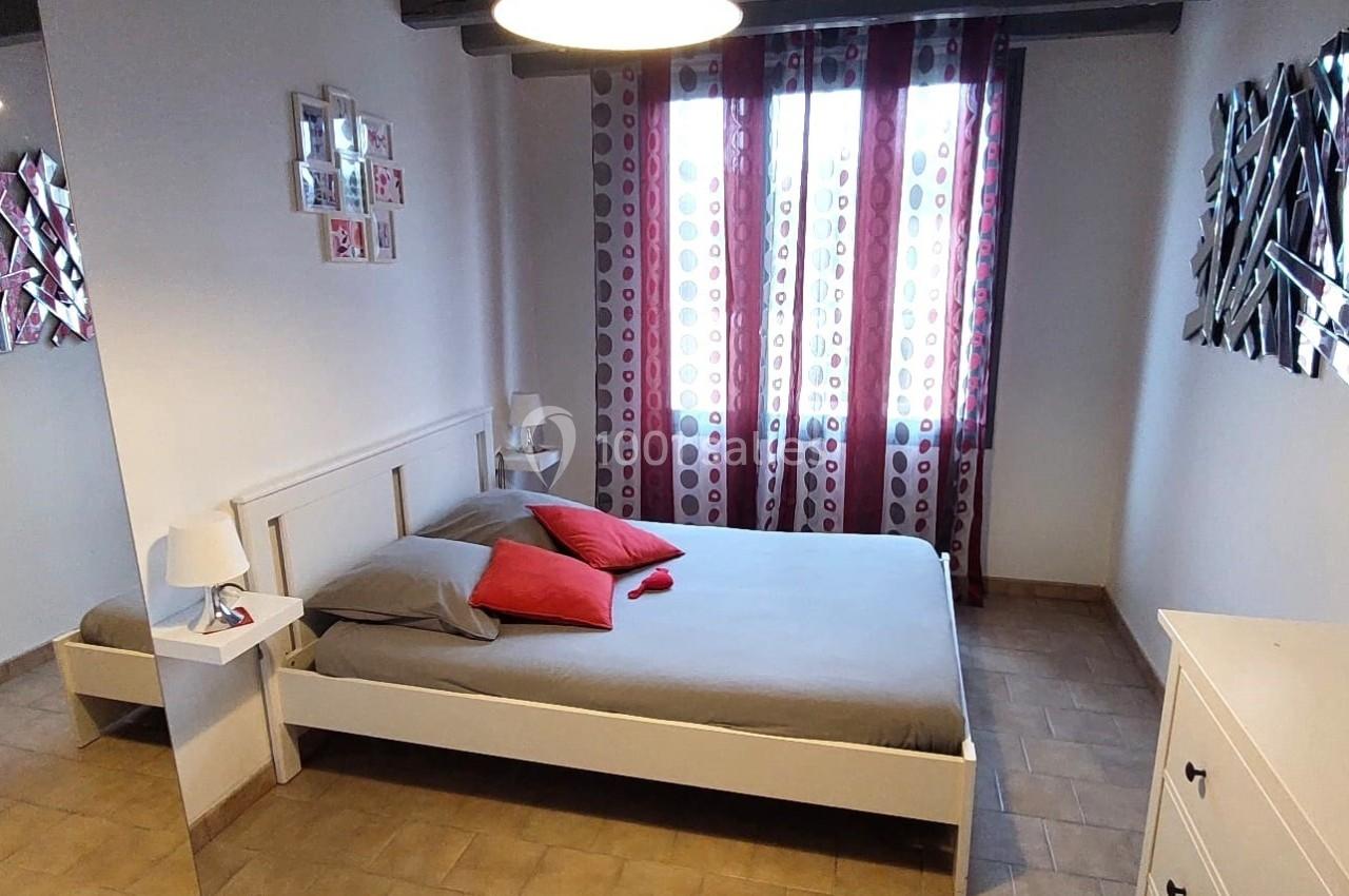 Chambre avec lit double, draps gris, coussins rouges, rideaux à motifs et éclairage suspendu.