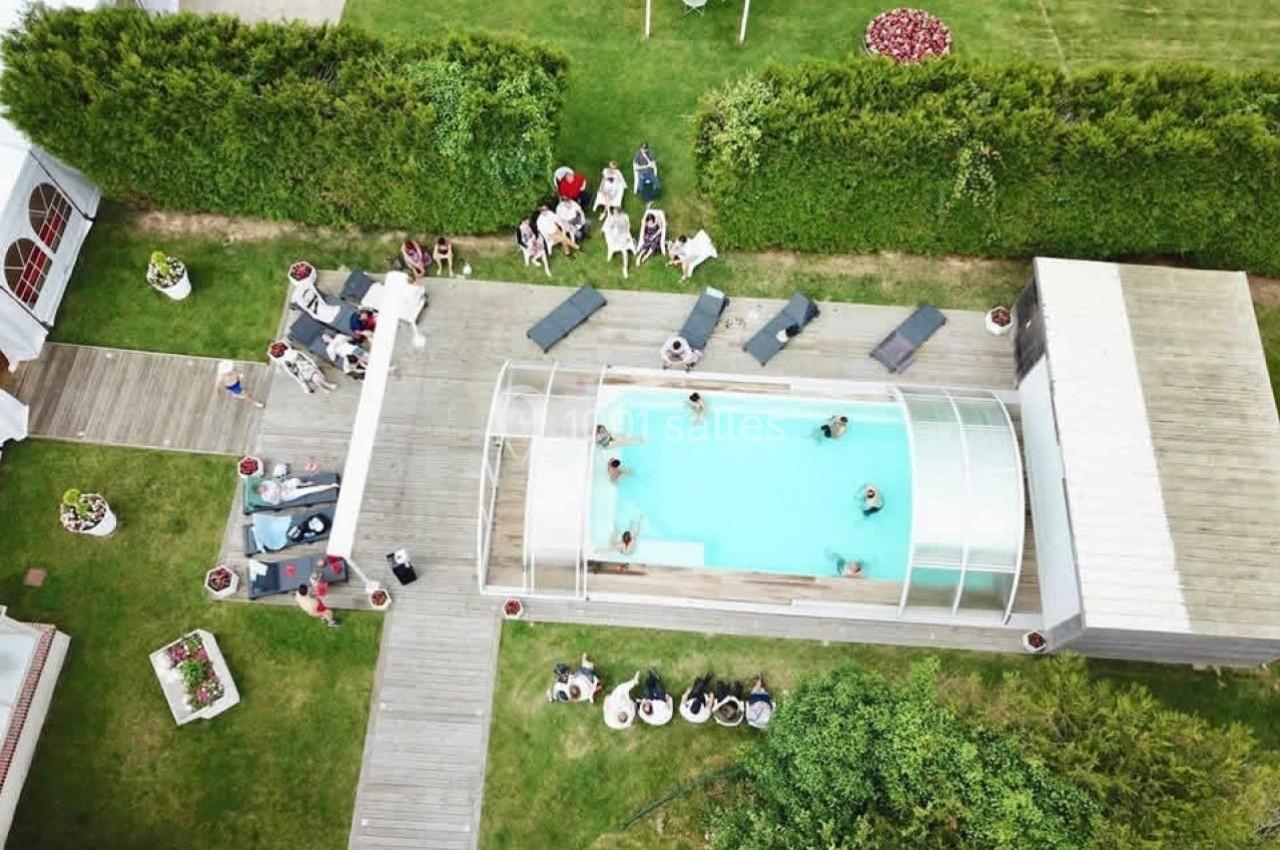 Vue aérienne d'une piscine extérieure couverte entourée de chaises longues, avec des personnes réunies dans un jardin.