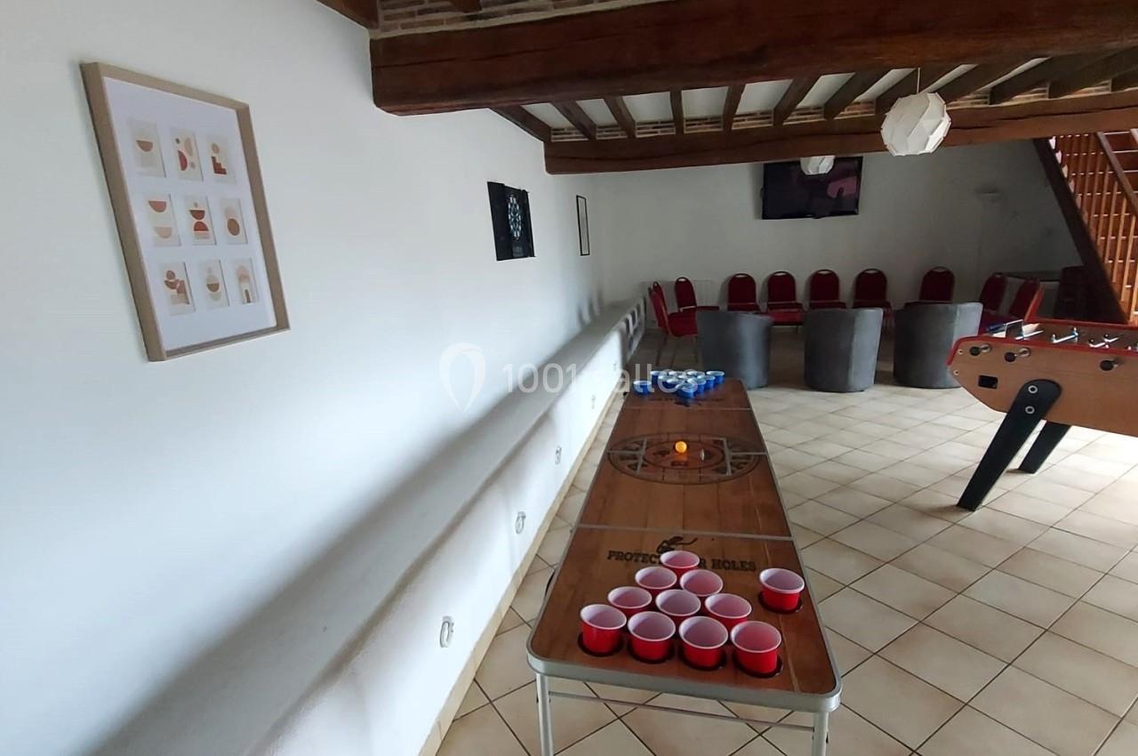 Table de beer-pong avec gobelets rouges dans une pièce à poutres apparentes, sol carrelé et baby-foot en arrière-plan.