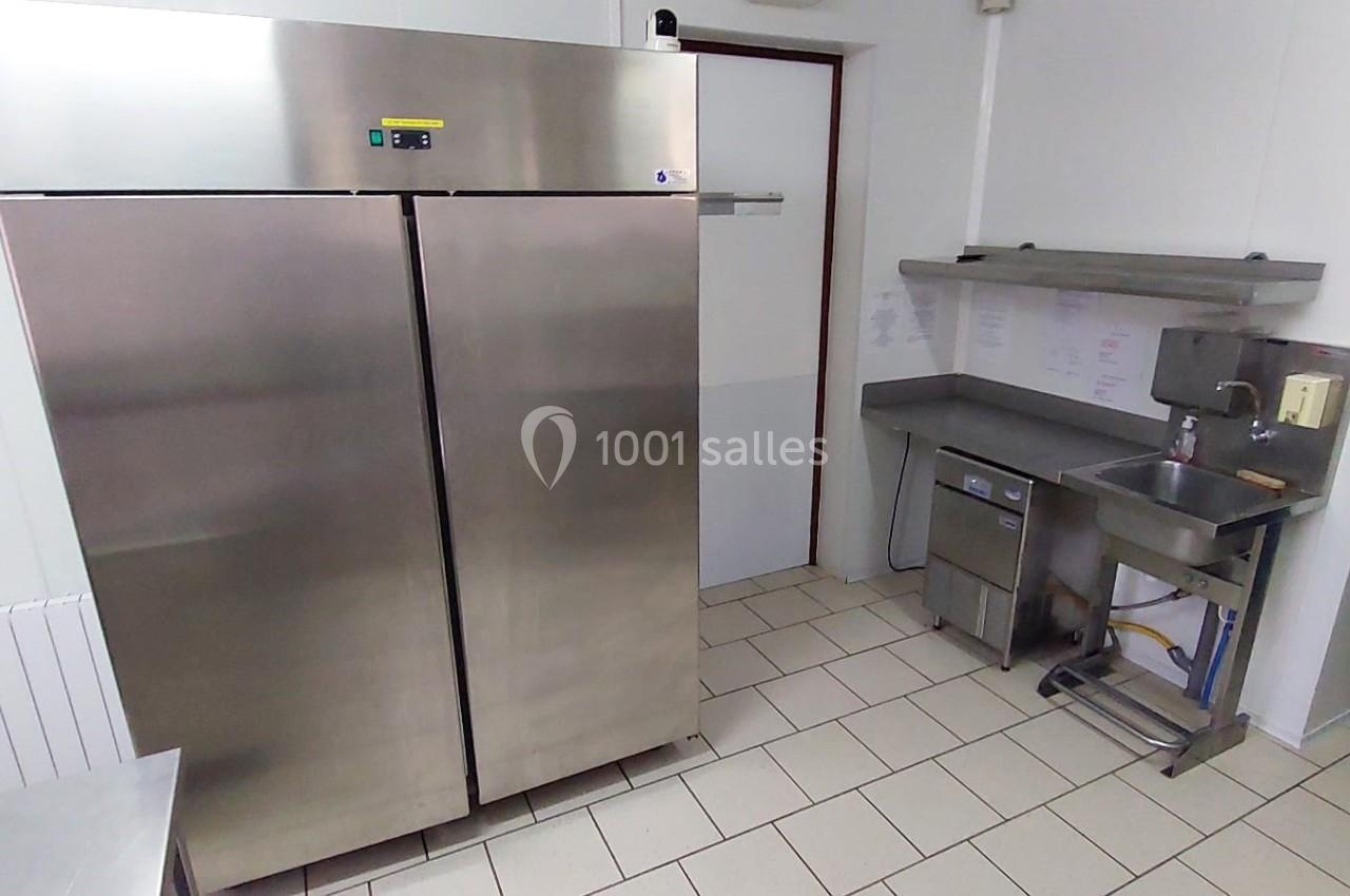 Cuisine professionnelle avec réfrigérateur en inox, évier, étagères murales et sol carrelé blanc.