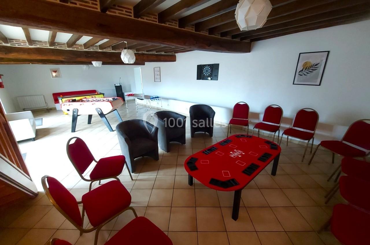 Salle avec des chaises rouges, fauteuils, tables de jeu, baby-foot et plafond avec poutres apparentes.