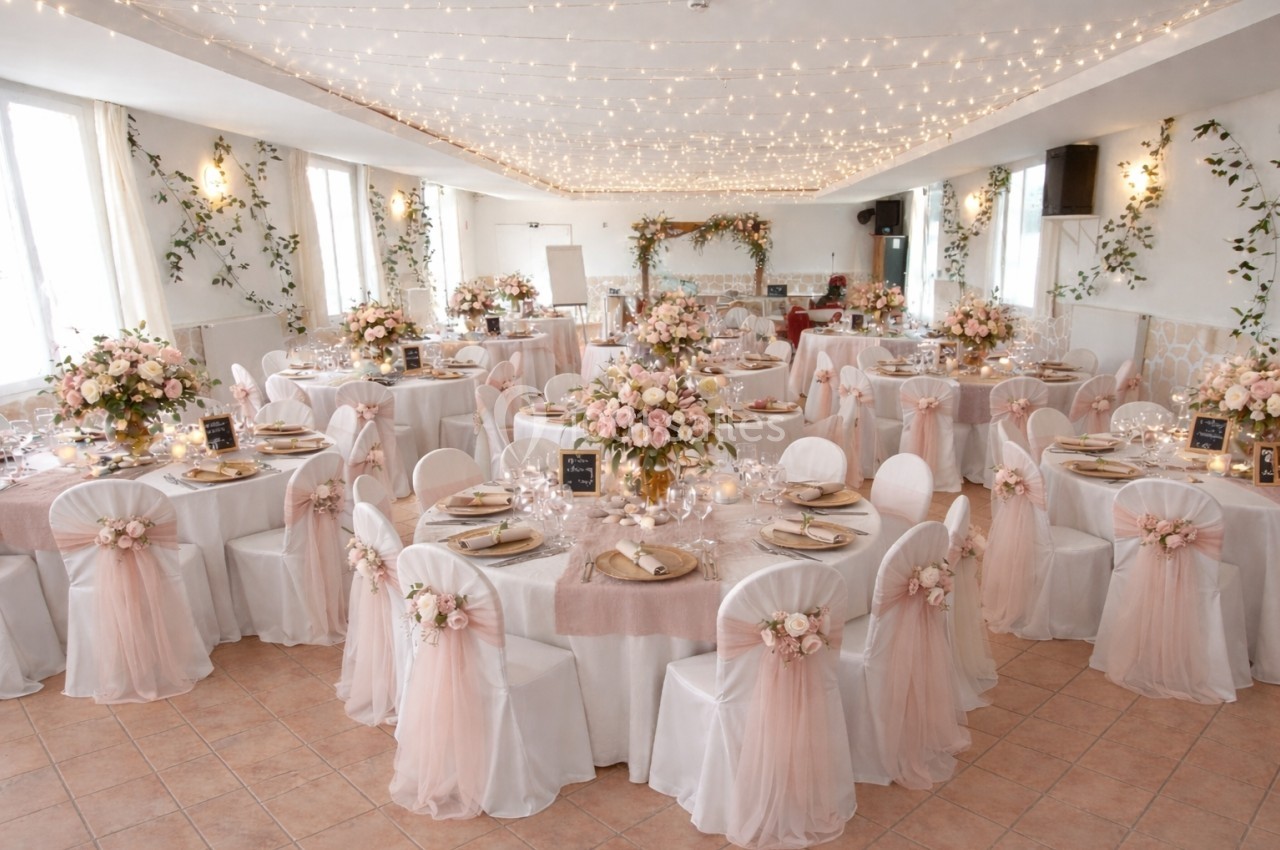 Salle de réception décorée pour un mariage avec tables rondes, nappes blanches, fleurs roses et guirlandes lumineuses.
