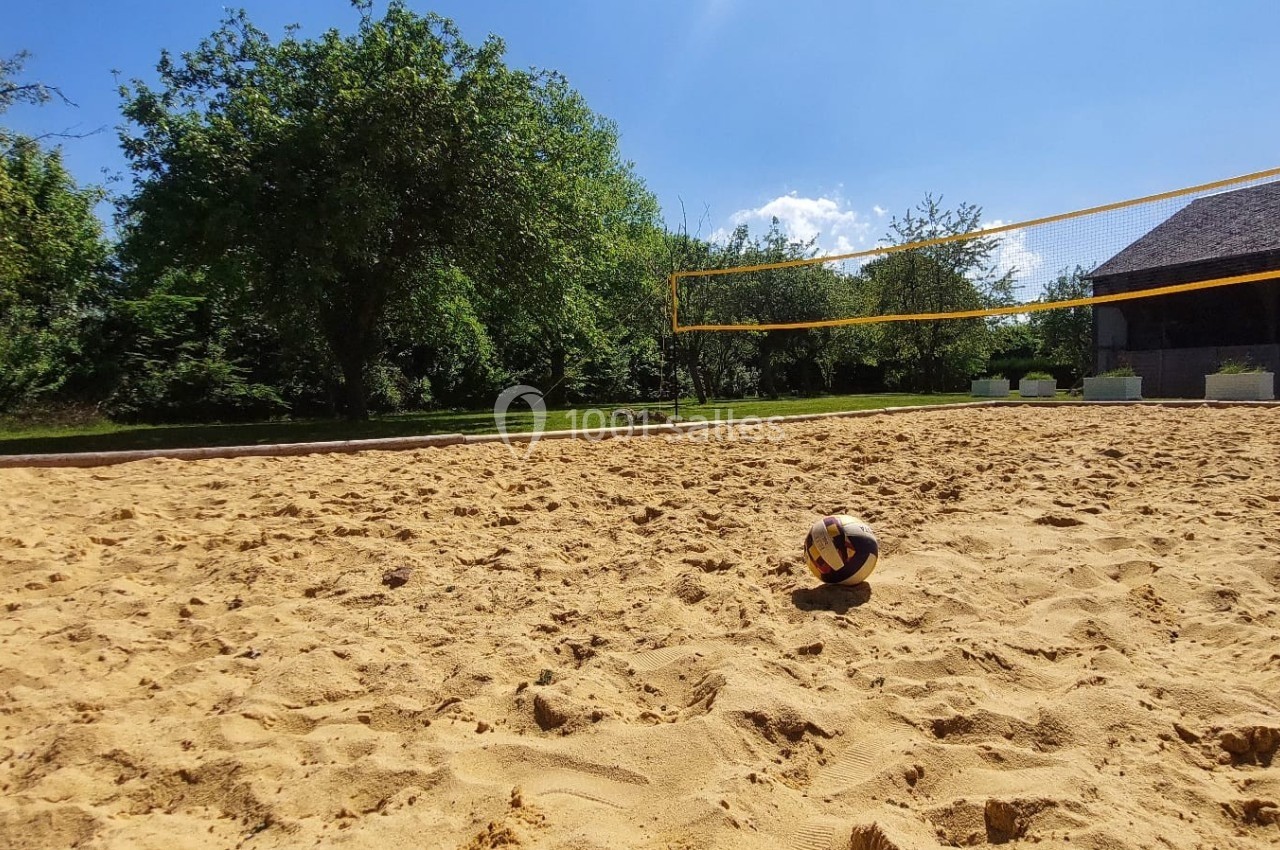 Terrain de beach-volley en sable avec un ballon au centre, entouré d'arbres et d'un bâtiment en arrière-plan.