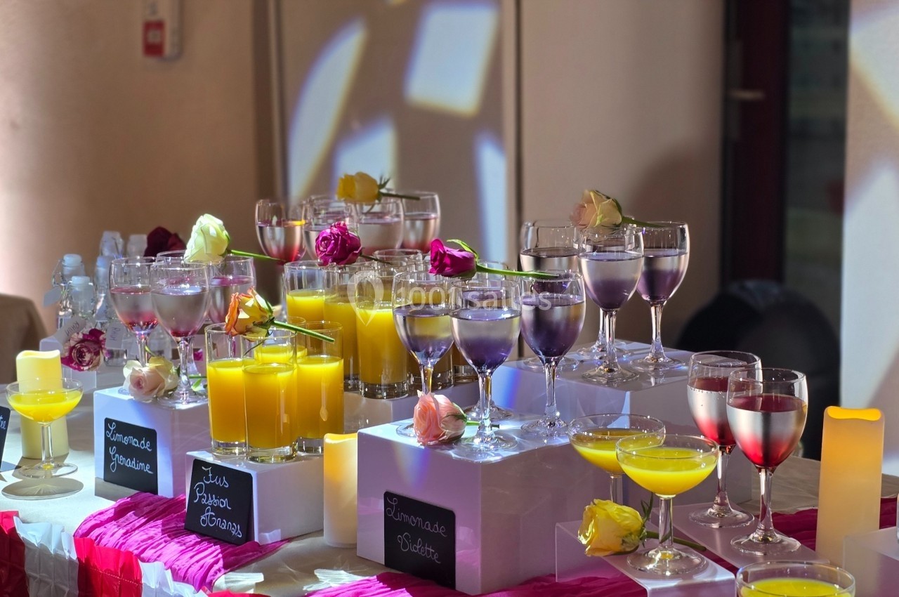 Buffet coloré avec verres de jus variés, étiquettes descriptives et décorations florales dans une salle éclairée.