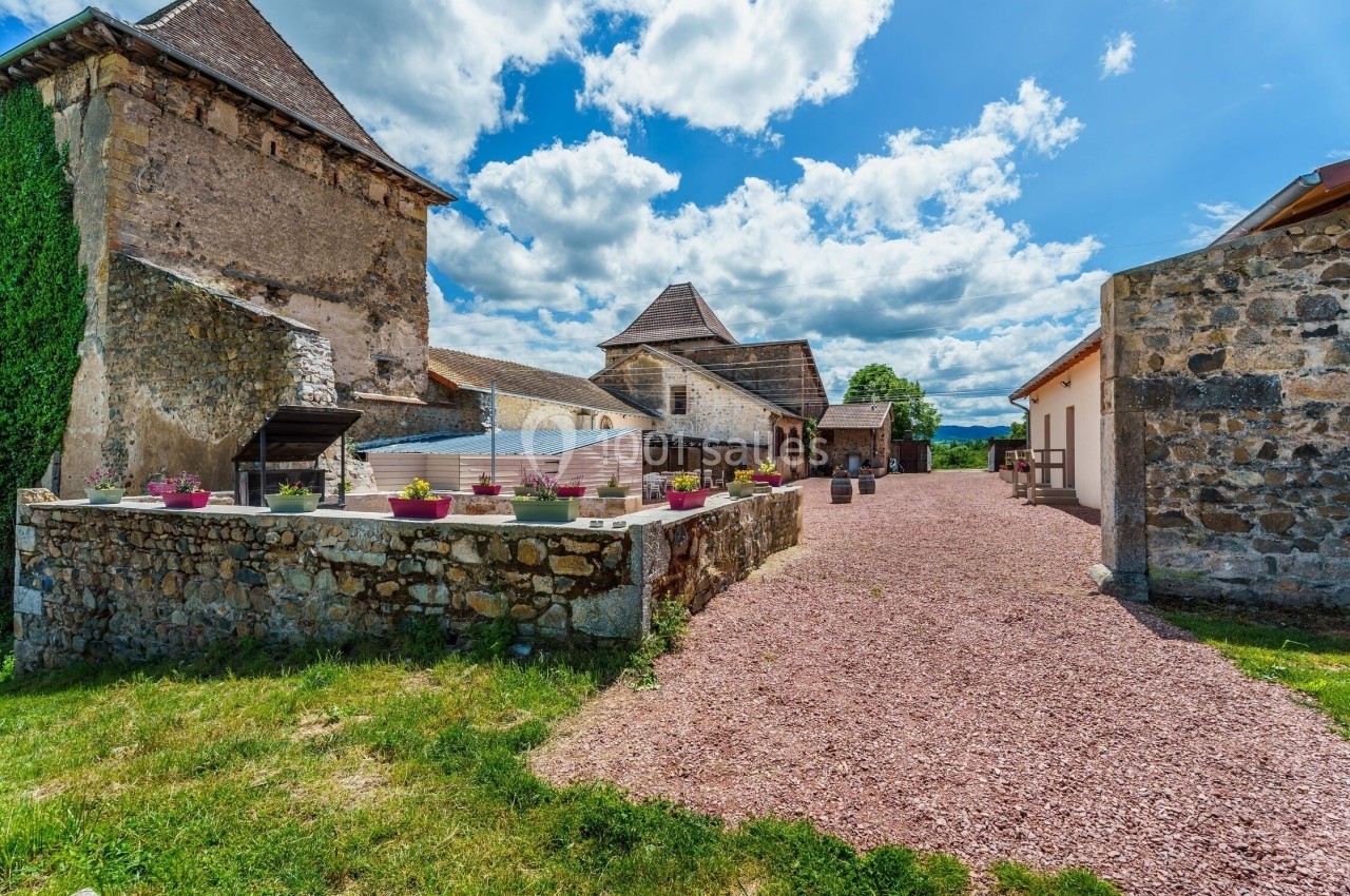 Cour d'une ferme ancienne avec bâtiments en pierre, terrasse fleurie et ciel partiellement nuageux en arrière-plan.
