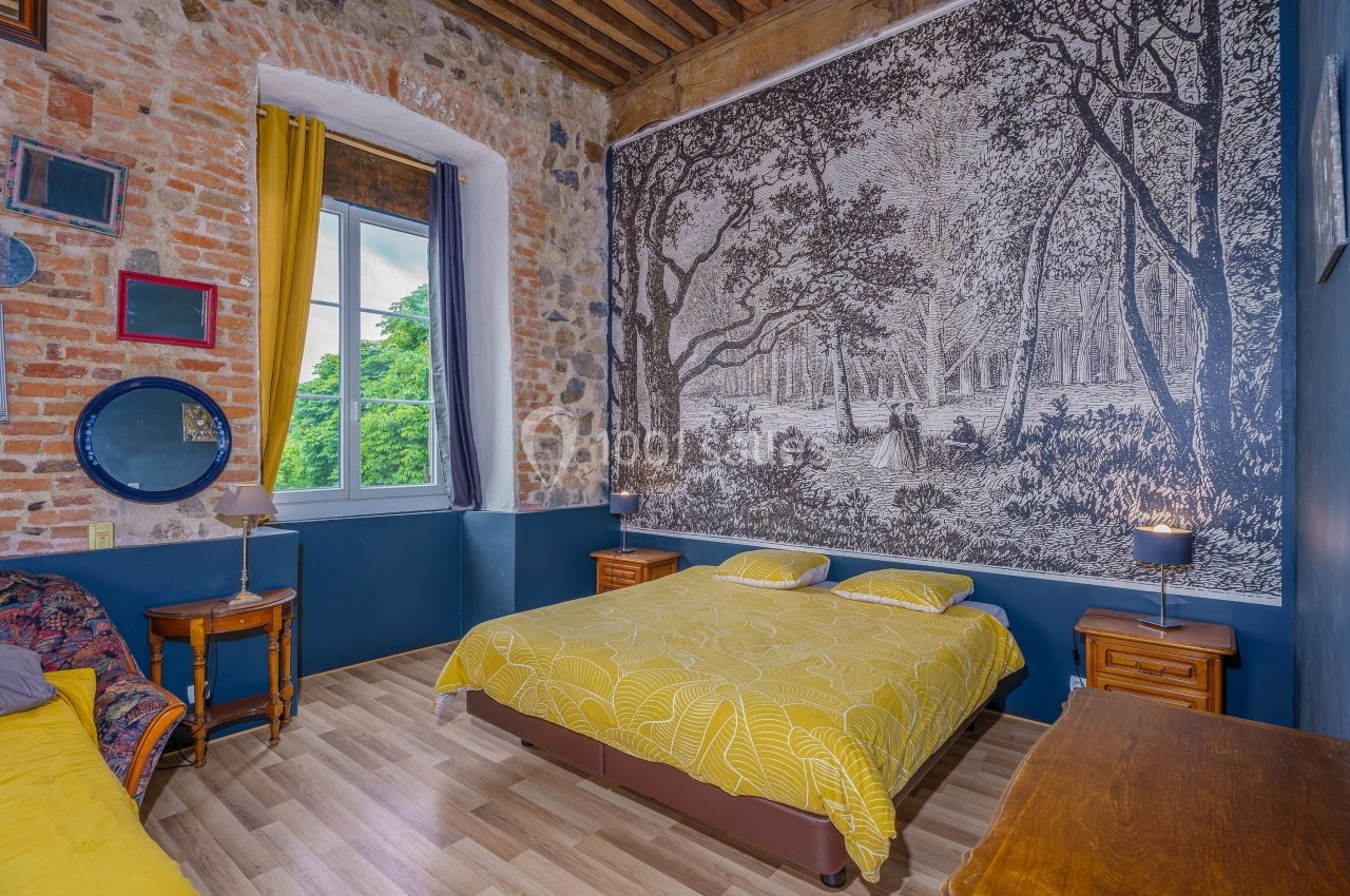 Chambre avec lit double, mur en pierre, grande tapisserie forestière, fenêtre lumineuse et mobilier en bois.