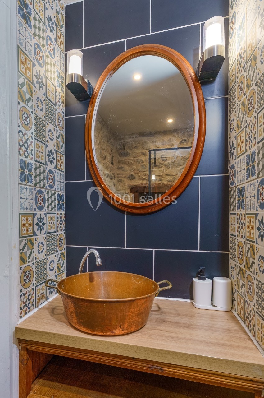 Lavabo en cuivre sur un plan en bois, miroir rond encadré de lumière, carrelage décoratif en arrière-plan.