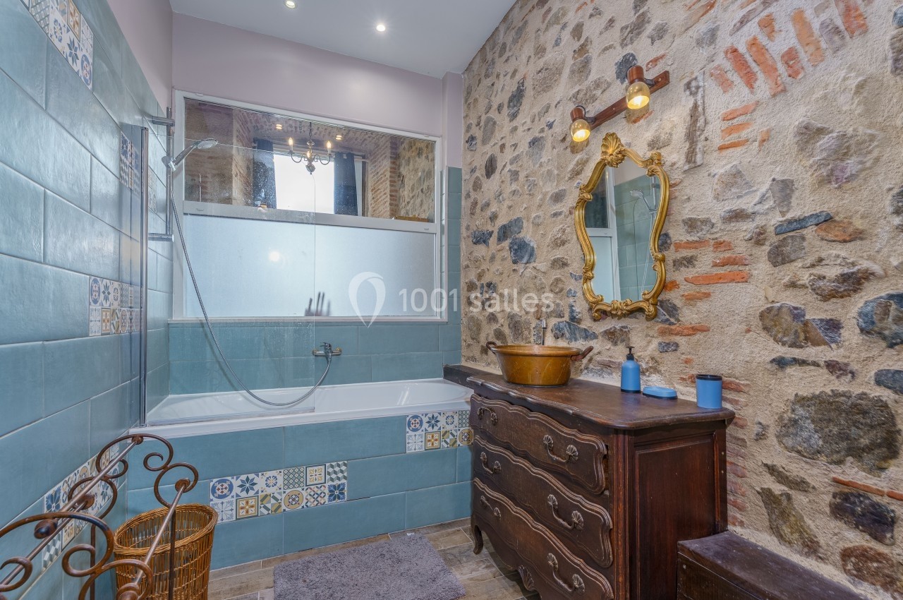 Salle de bain avec mur en pierre, baignoire à carreaux bleus, meuble ancien en bois et miroir doré.